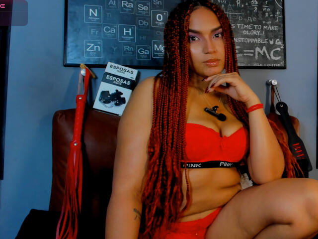 BrianaPratt with red braided hair, red Victoria's Secret lingerie, showing her tits, sitting on a brown leather chair, looking at the camera with a slight smile.