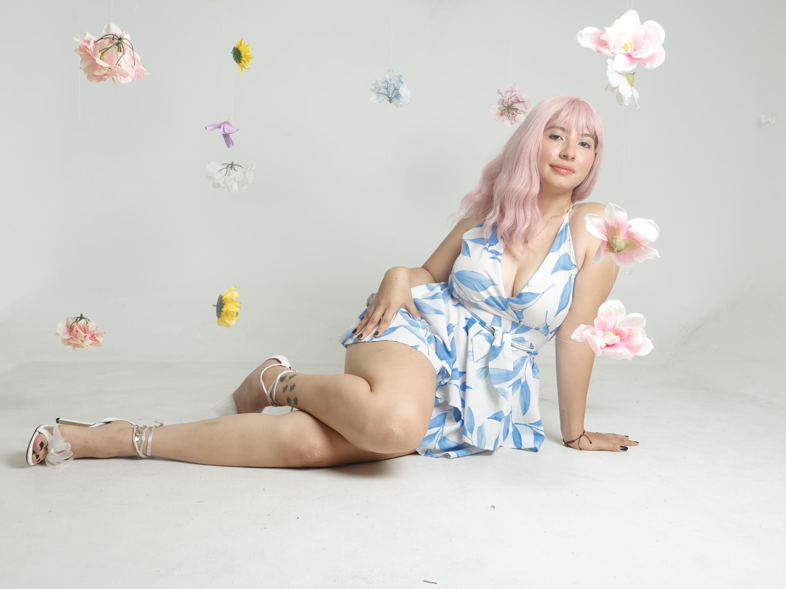 EmmaWren with pink hair, wearing a blue floral dress, white heels, and no panties, showing her tits and pussy, surrounded by floating flowers, laying on a white floor.