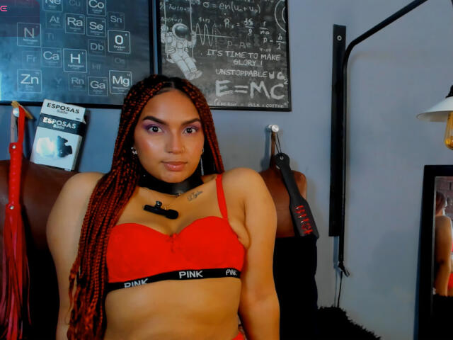 BrianaPratt with red bra, black choker, and braids, looking at camera. She has small tits, light brown skin, and a tattoo above her cleavage. Background is a gray room.