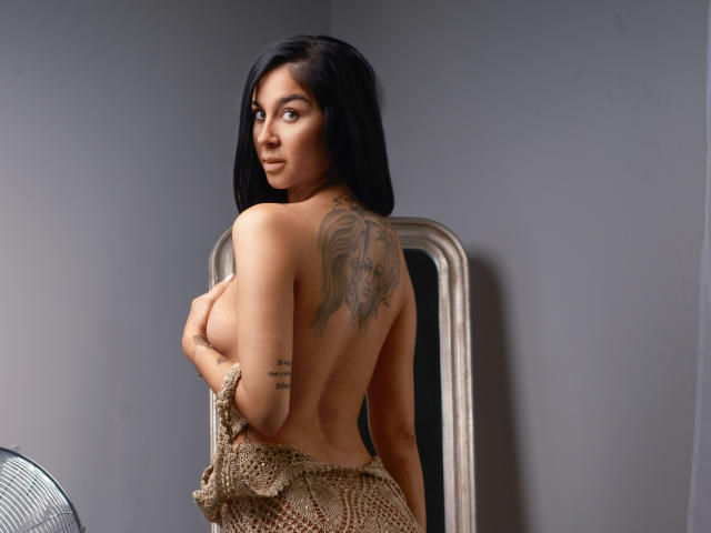 FrancescaDante with long black hair, tan skin, and a large back tattoo, topless, covering her tits with a beige netted cloth, standing in a gray-walled room.