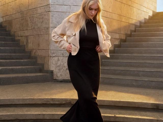 AlinaLemon tiene el cabello rubio, lleva una chaqueta beige sobre un vestido negro de estilo sirena, está de pie en escaleras de piedra con las manos en las caderas, mirando hacia abajo..