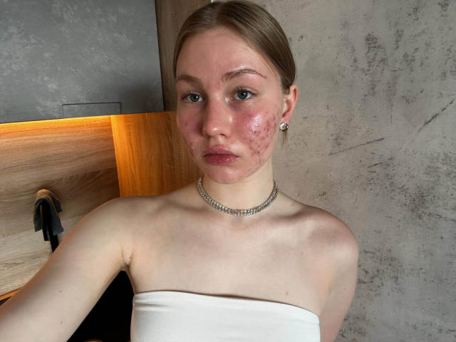 ZofiaZhu with puffy, red-assed cheeks, wearing a white strapless top, silver choker, and earrings, in a wooden and concrete room, looking straight at the camera.