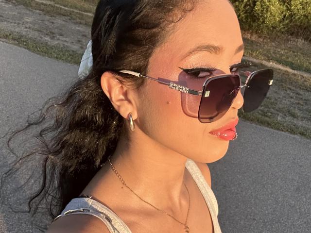 NashiroSakuraii with glossy red lips, dark wavy hair in a white scrunchie, wearing oversized Ray-Ban sunglasses, gold hoop earrings, and a white tank top, sunlight on her face.