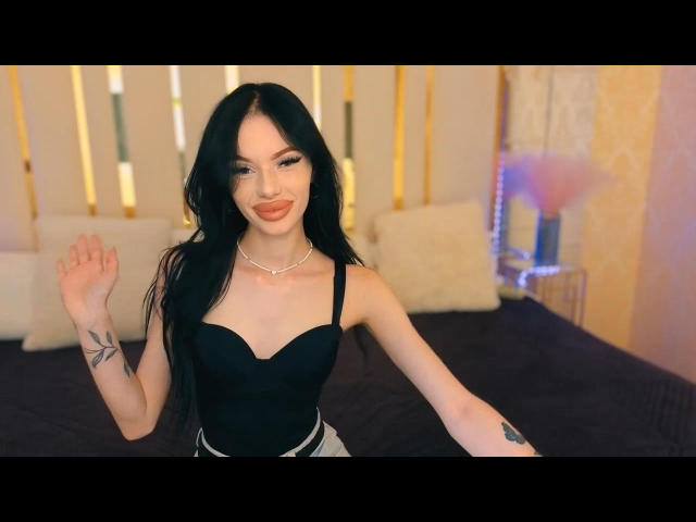 SamantaBredShow with long black hair, wearing a black strapless top, showing her small tits, a tattoo on her left arm, and a necklace, smiling with her right hand up.