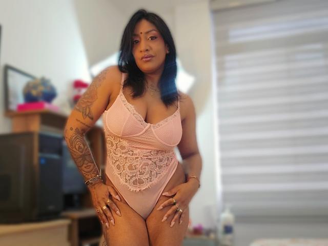 Nahirr, curvy Latina with black hair, wearing a pink lace lingerie bodysuit, showing her large tits and ass, standing in a bedroom with tattoos on her arm.