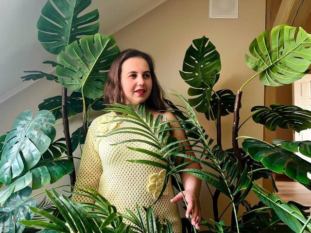 DariaCalm with medium tits, wearing a yellow polka-dot dress, stands amidst large green monstera leaves, smiling softly, brown wavy hair, light skin, in a sunlit room.