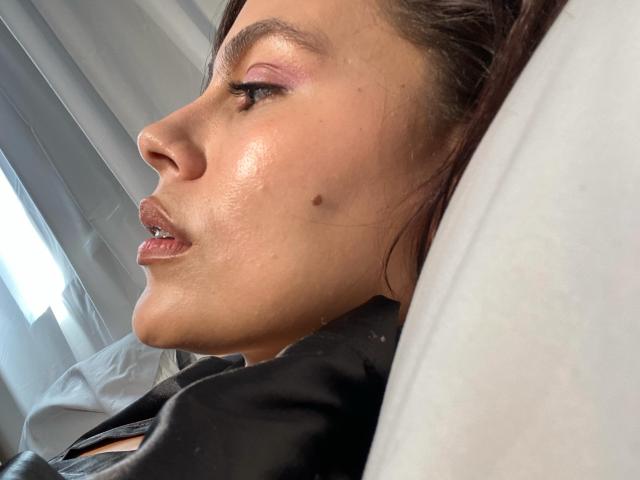 El rostro de RossaHot en un perfil lateral, mostrando una pequeña marca de nacimiento en su mejilla, con sombra de ojos rosa y labios brillantes, acostada sobre un almohadón blanco..