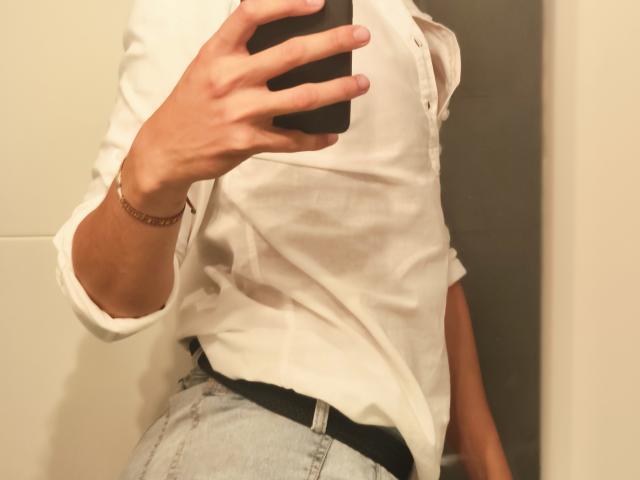 Teronn draagt een wit overhemd met knopen, houdt een telefoon vast, met een zichtbare armband om zijn arm en pols, draagt lichtblauwe jeans en een zwarte riem, spiegelzelfportret..