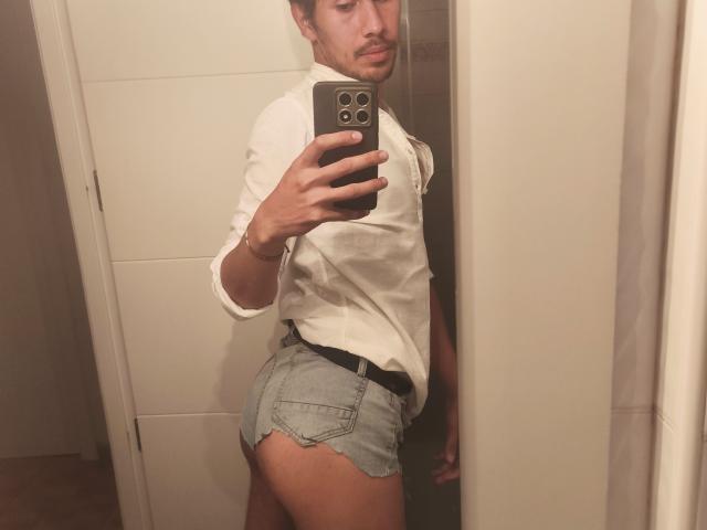 Teronn with a white shirt and tight, frayed denim shorts, showing his ass in a mirror selfie, holding a black phone with three cameras.
