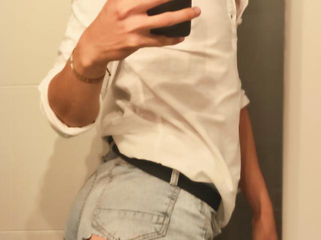 La camisa blanca de Teronn, los jeans azules ajustados y el cinturón negro, con una pulsera dorada en su muñeca derecha, se toma un selfie con un espejo. En su mano sostiene un teléfono negro..