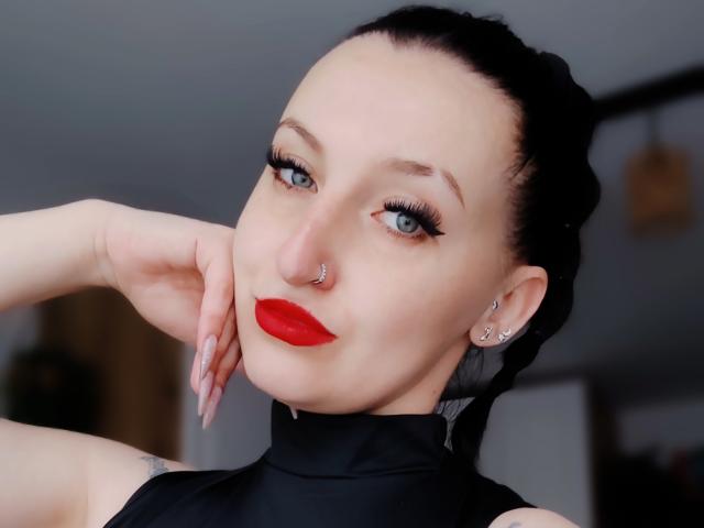 GoddessMoonx hat blasse Haut, blaue Augen, roten Lippenstift, eine schwarze Nasenring und schwarzes Haar, das zu einem Pferdeschwanz gebunden ist. Sie trägt ein schwarzes Top ohne Ärmel und hat eine Hand an ihrer Wange..