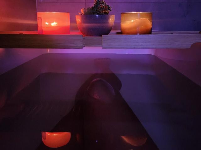 Aurora Monroes Beine in einer dunklen, violett beleuchteten Badewanne, mit drei leuchtenden Kerzen auf einem hölzernen Regal darüber, die orange- und rot gefärbtes Kerzenlicht reflektieren. Eine Topfpflanze mit Sukkulenten in der Mitte..