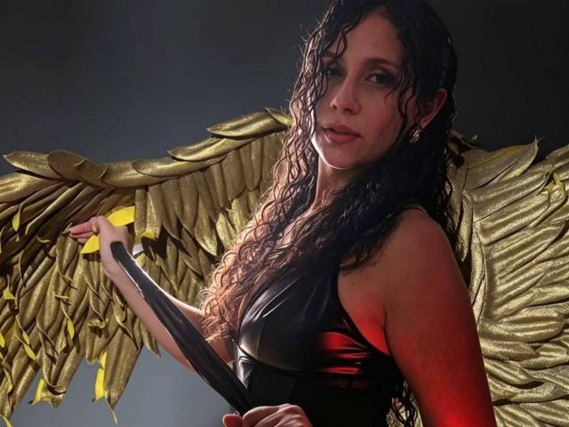 RoseeCarolina with wet curly black hair, golden wings, and a black shiny top, holding a black strip, looking slightly to the side with a serious expression.