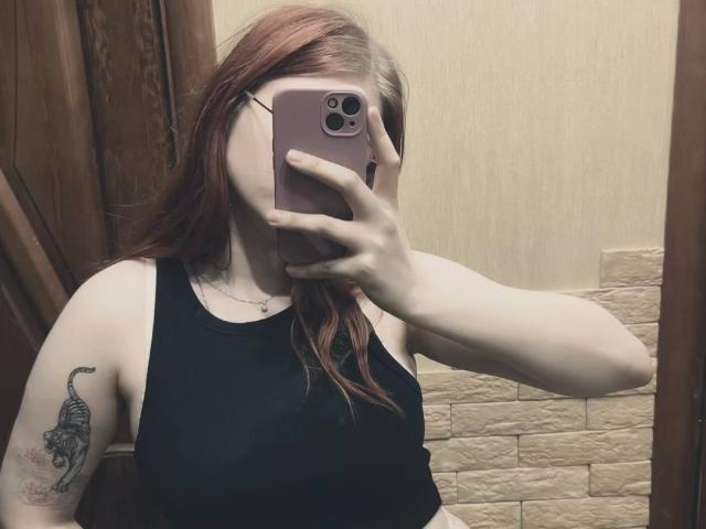 MargSeyler with red hair, black tank top, holding pink phone, covering face, tattoo of scorpion on right arm, in mirror selfie, showing pale skin and small tits.