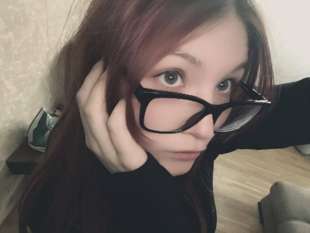 MargSeyler with red hair, black glasses, pale skin, and blue eyes, holding her hair, wearing a black top, in a selfie, showing her face and hand.