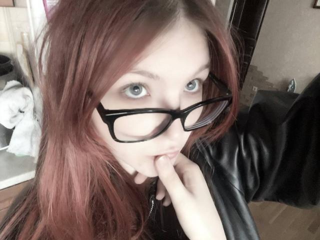 MargSeyler with red hair, black glasses, sucking her finger, pale skin, blue eyes, black leather jacket, in a selfie, showing her mouth and finger.