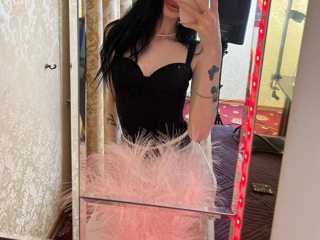 SamantaBredShow in a black strapless corset, showing her tits, with long black hair, red lipstick, and tattoos, taking a mirror selfie, with pink furry trim in foreground.