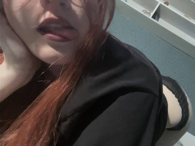 MargSeyler has red hair, black top, and black panties, on all fours with tongue out, showing her ass and white skin. Her hand's on the side of her face.