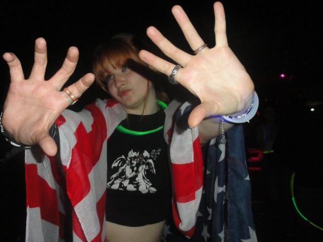 MargSeyler with pale skin, brown hair, and bangs, wearing a red and white striped jacket, black shirt, and rings, hands up in front, looking at camera.