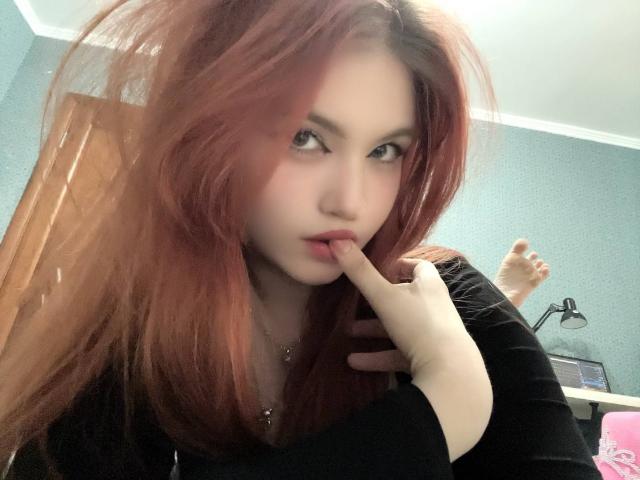 MargSeyler with red hair, biting her finger, wearing a black top, light skin, and a subtle nose piercing, in a slightly blurred selfie.