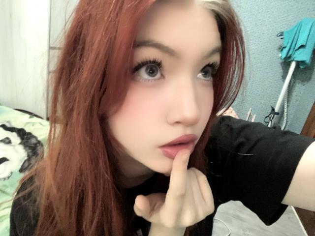 MargSeyler has pale skin, red hair, and blue eyes, with a finger on her pink lips, wearing a black shirt, in a selfie. Her eyelashes are long and dark.