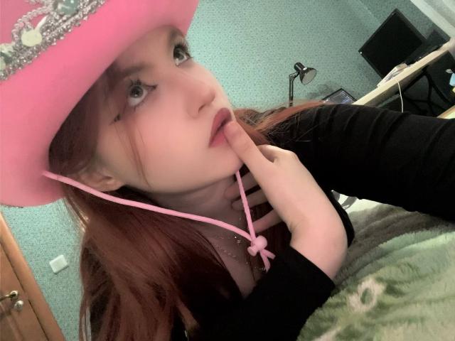 MargSeyler with red hair, pink hat, and black top, sucking on her finger with pink earbuds, showing her tits under a green blanket.