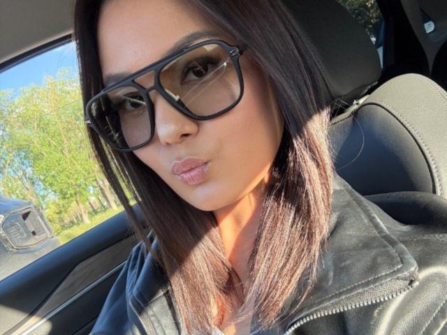 Johana Urlanza, con cabello castaño oscuro, usando gafas negras y una chaqueta de cuero negra, frunciendo sus labios carnosos, sentada en un coche con la luz del sol en su rostro..