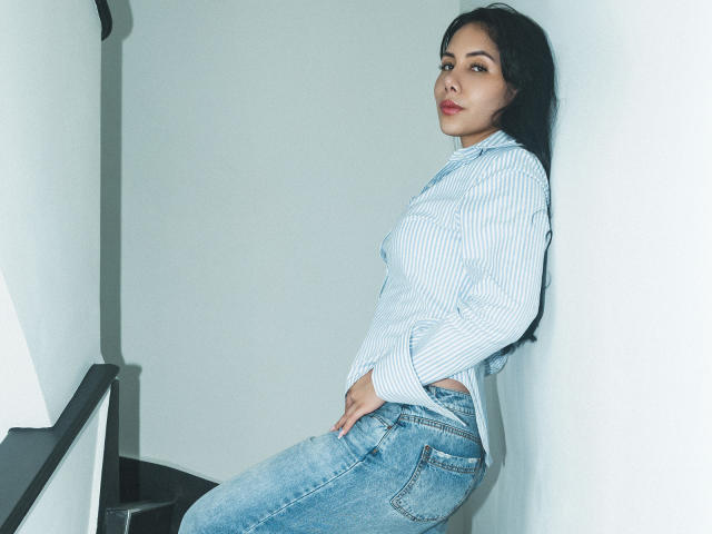 ArianaRogers leaning against a wall, wearing a blue striped shirt and blue jeans, with her hand on her ass, dark hair, red lipstick, and a confident look.