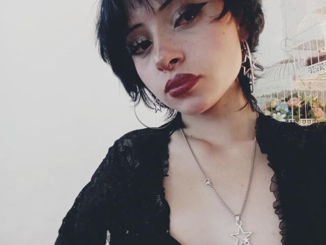 ViolettClay with short black hair, dark eyeliner, red lipstick, nose ring, black lace top, and star necklace, showing small tits, looking at camera, in a soft-lit setting.