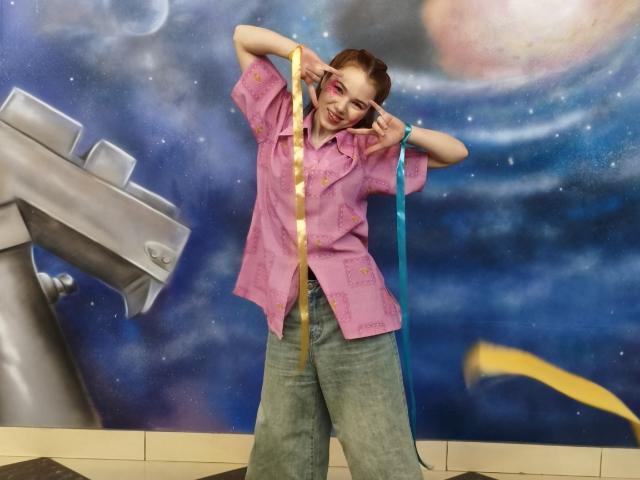 KatiRokisky con una camisa rosa, jeans azules, sosteniendo cintas amarillas y azules, con los brazos levantados, haciendo el gesto de "rock", sonriendo, cabello castaño, sombra de ojos rosa, fondo con temática espacial..