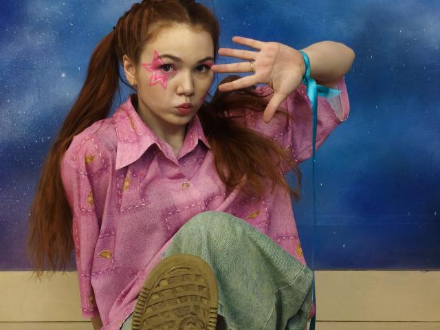 Kati Rokisky mit pinkem Stern-Make-up, einem pinken Hemd mit Knopfleiste, grünen Hosen und braunen Schuhen, die ein Friedenszeichen zeigt, mit blauem Band, braunen Zöpfen und einem schmollenden Gesichtsausdruck..