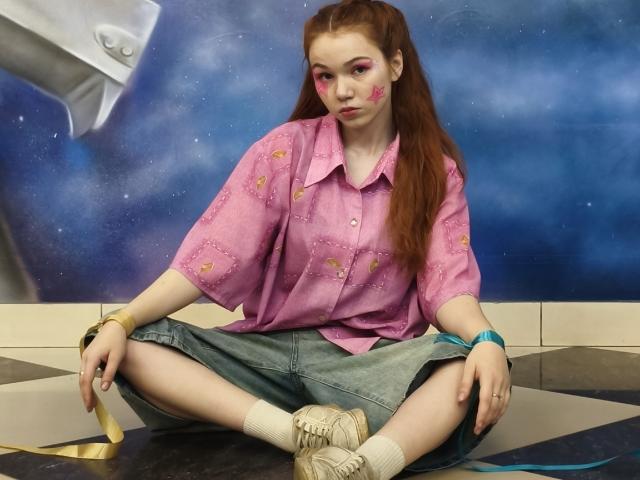 KatiRokisky, con cabello rojo, pintura rosa en la cara, una camisa rosa, pantalones cortos vaqueros, calcetines blancos y zapatos beige, sentada con las piernas cruzadas sobre un suelo a cuadros, y con pulseras amarillas y azules..