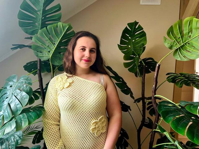 DariaCalm tem seios de tamanho médio, vestindo um vestido amarelo de malha com bordados florais, e está em frente a grandes folhas de monstera verde, sorrindo suavemente, com cabelos castanhos ondulados..