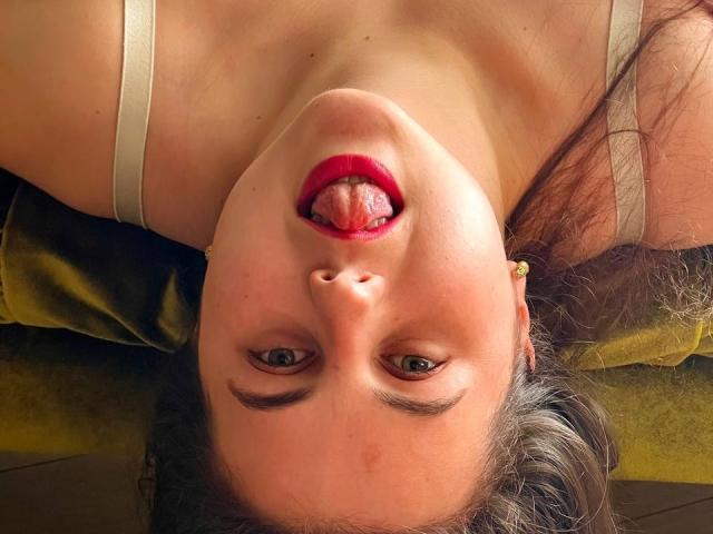 Il viso di DariaCalm è capovolto, con la lingua fuori, rossetto rosso, occhi verdi e capelli castani, indossa una maglietta beige e giace su una superficie giallo-verde..