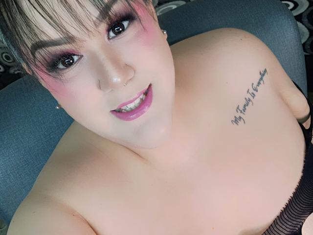 GiiannaMorrys has pink eyeshadow, a nose piercing, and a tattoo on her right shoulder. She's smiling, showing her teeth, with her bare shoulders and black top visible.