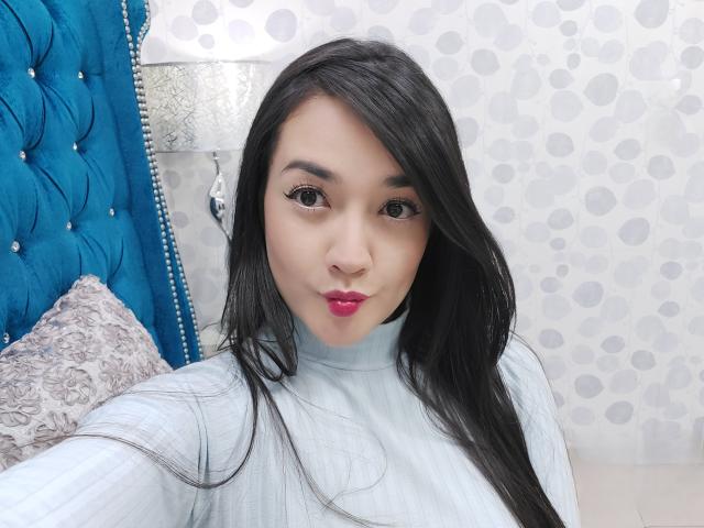 Annhy with long black hair, red lipstick, and a light blue turtleneck, sitting in front of a blue tufted headboard, looking at the camera with a slightly pouty expression.