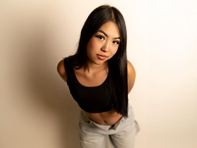 Jules Durand hat schwarzes, glattes Haar, braune Augen und eine kleine Nase. Sie trägt ein schwarzes Crop-Top und hellgraue Hosen und blickt mit leicht zusammengekniffenen Lippen nach oben..