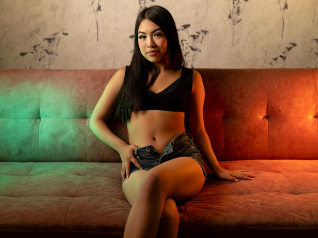 Jules Durand, con cabello negro largo, vestida con un top corto negro y pantalones cortos vaqueros, sentada en un sofá de terciopelo iluminado en tonos verde y naranja, con una mano en la ingle y las piernas cruzadas..