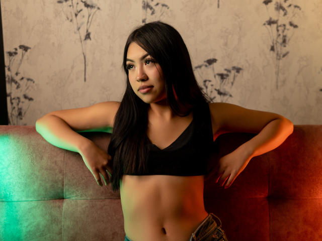 JulesDurand with long black hair, wearing a black sports bra, leaning on a red couch, green and orange light reflecting on her, hands on her hips, looking at camera.