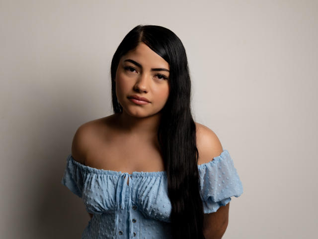 Briana Clark X mit langen, schwarzen Haaren, heller Hautfarbe, trägt ein blaues Kleid mit einem Schulterteil, das die Brust betont, und hat einen dezenten, selbstbewussten Gesichtsausdruck, vor einem schlichten Hintergrund..