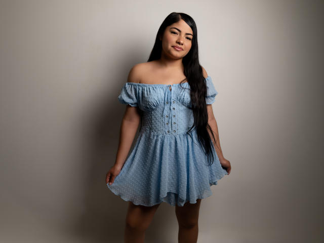 BrianaClarkX, con cabello negro largo, vestida con un vestido azul de un solo hombro, sosteniendo la falda, con botones en la parte delantera, con senos pequeños, una ligera sonrisa, de pie frente a una pared lisa..