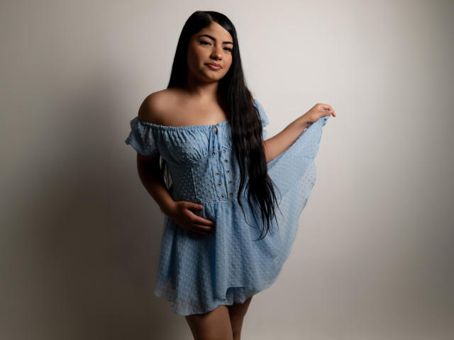 BrianaClarkX met lang zwart haar, gekleed in een blauwe jurk met een open schouder, die de rok vasthoudt, lachend, waarbij haar borsten en billen zichtbaar zijn, staand voor een effen achtergrond..