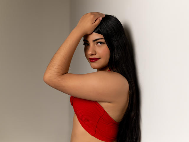 BrianaClarkX mit langen, schwarzen Haaren, rotem BH und einem Arm, der den Kopf bedeckt, lächelt mit rotem Lippenstift, zeigt glatte, gebräunte Haut und kleine Brüste, und steht vor einer weißen Wand..