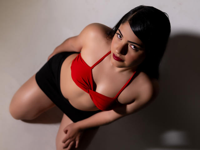 BrianaClarkX, avec ses cheveux foncés, son soutien-gorge rouge et son short noir, regarde vers le haut avec du rouge à lèvres, de larges seins et des hanches généreuses, agenouillée sur une surface claire..