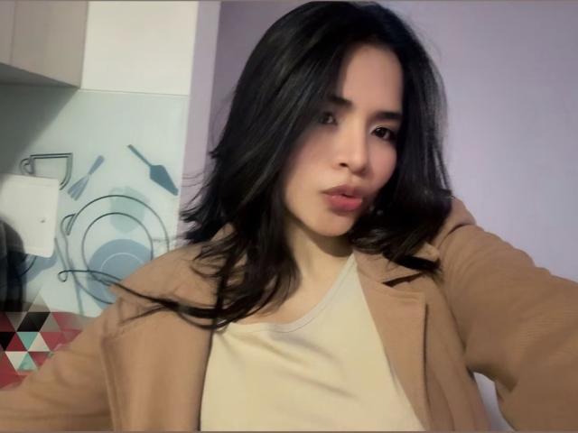 SaschaHelen, con lunghi capelli neri, labbra piene, indossa un cappotto beige sopra una maglietta bianca, in una cucina con una parete a motivi. Il seno non è visibile, l'attenzione è concentrata sul viso..