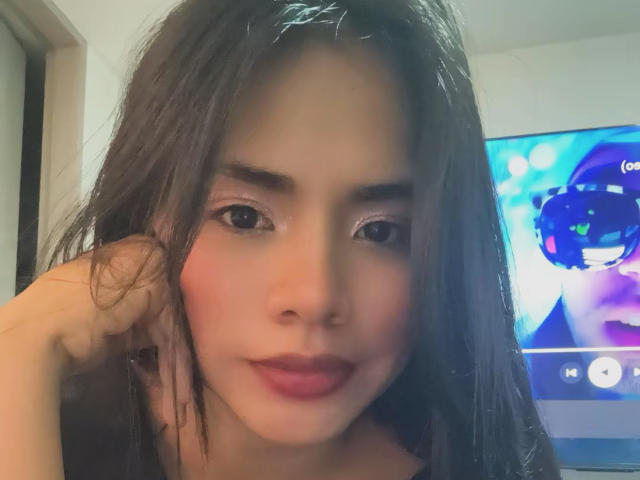 SaschaHelen has wet black hair, pinkish blush on cheeks, dark eyebrows, and pink lipstick. She's touching her hair with a slight smile, and a TV is in the background.