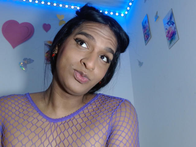 IdiiaLatina with dark hair in buns, purple fishnet top, pouty lips, and cute expression, showing small tits through mesh, looking to the side with blue LED lights.
