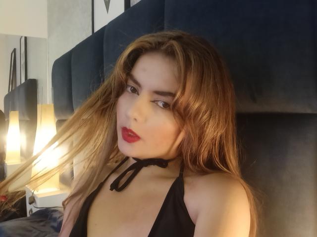 MandyMouses con lunghi capelli castani, rossetto rosso, top nero con spalline e un leggero sorriso, che si toccano i capelli, su una testiera blu in velluto, con un letto e lampade sullo sfondo..