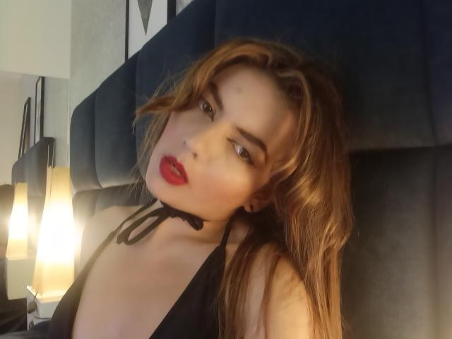 MandyMouses met rode lippen, een zwart haltertopje en bruin haar, leunend tegen een donkerblauwe hoofdbord, kijkend naar de camera met een licht geopende mond..