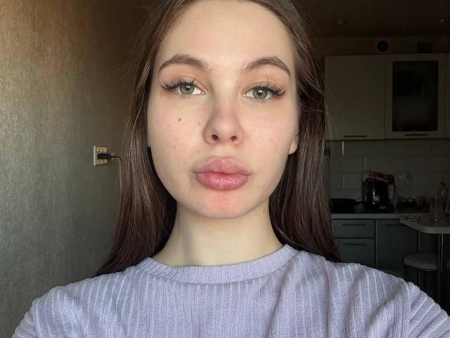 KarleenTallas with pale skin, green eyes, and brown hair wearing a purple rib-knit sweater, puckering her lips in a selfie. Her tits are small, and her ass is unseen.